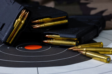 Loaded rifle magazines with loose rounds of ammunition scattered on top of a paper target with a camouflage background. Background and foreground blurred with a shallow depth of focus.