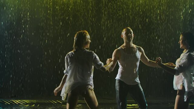 Passionate movements of a salsa dance in the rain on a dark smoky backlit background. A silhouette of three wet bodies moving seductively in slow motion. Close up.