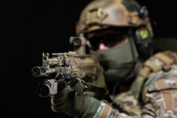 Close up of a soldier in camouflage and full military gear, wearing a helmet, glasses and protective mask, raises a rifle to his face and taking aim, preparing to shoot, isolated on black background