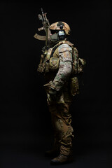 Full length side view portrait of an army soldier standing in full military uniform, wearing a bulletproof vest, helmet, glasses and mask, holding up a submachine gun isolated on a black background