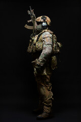 Fototapeta premium Full length side view portrait of an army soldier standing in full military uniform, wearing a bulletproof vest, helmet, glasses and mask, holding up a submachine gun isolated on a black background