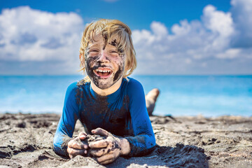 11.11 Black Friday concept. Smiling boy with dirty Black face sitting and playing on black sand beach. Summer vacation with kids. Black Friday, sales of tours and airline tickets or goods
