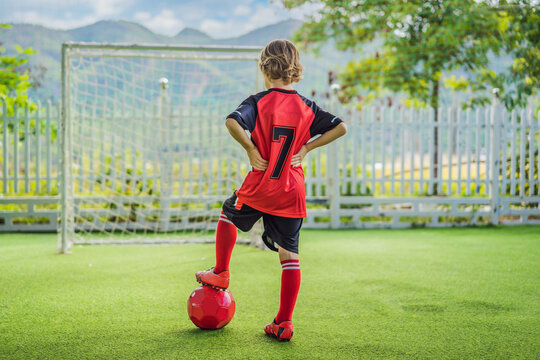 Little Cute Kid Boy In Red Football Uniform Playing Soccer, Football On Field, Outdoors. Active Child Making Sports With Kids Or Father, Smiling Happy Boy Having Fun In Summer