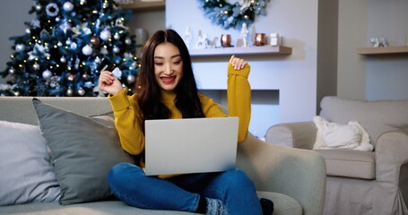 Happy Asian smiling woman sitting at home in decorated room on Christmas Eve and buying xmas gifts online on laptop paying with credit card. Holiday shopping. Xmas discounts. E-commerce concept