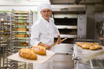 male baker with bread on baking shovel in kitchen - obrazy, fototapety, plakaty