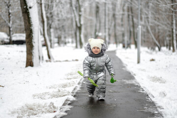 Fototapeta na wymiar Happy child on a winter walk in the park. A funny girl in a warm silver overalls runs along the path. The baby is holding a toy shovel. First snowfall. Blurred background.