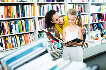 Portrait of happy young woman with girl reading textbook in book shop. Focus on woman
