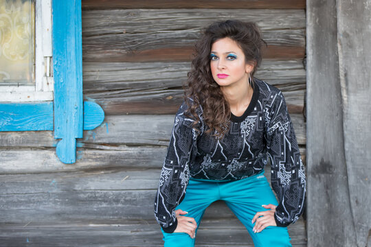A brunette is standing against a wooden wall, dressed in the style of the nineties with bright makeup. A funny woman in sweatpants with beautiful makeup.
