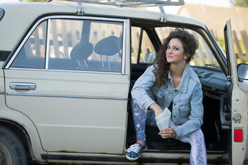 Amusing young woman in a denim jacket with a bag of sunflower seeds sits in an old car. Woman in the style of the eighties.