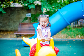 A cheerful girl with two pigtails on her head is riding a swing in the park and laughing. Emotional portrait of a child in the park