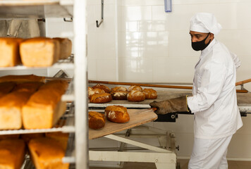 Baker of small bakery in face mask for viral protection holding hot bread on shovel just from oven