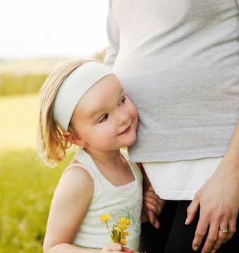 Little girl holding a handful of buttercups and resting her head on mother's pregnant belly listening ror baby