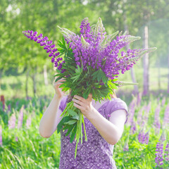 A young woman with a large bouquet of purple lupine flowers stands in a meadow.
