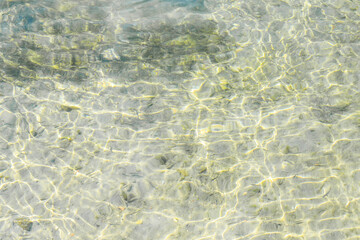 Shallow clear green and yellow water with algae and small stones on the bottom. Shiny light reflections pattern and waves