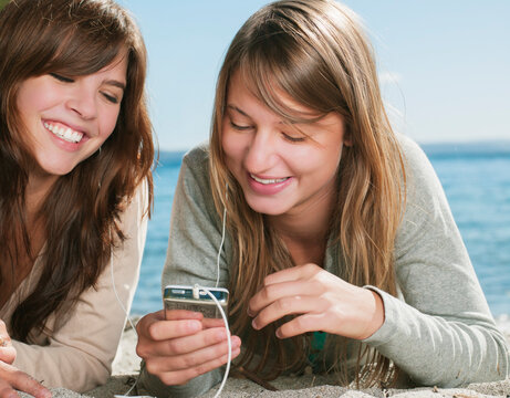 Two teenage girls laying on the sand, sharing earbuds, listening to music