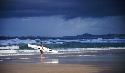 Side on view of female surf lifeguard training walking along the beach holding ocean surf ski with rough waves and stormy sky