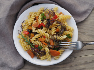 Pasta with a sauce of different vegetables and herbs in a plate on a wooden background, top view. Healthy dish with curly macaroni and stewed vegetables for a proper vegetarian diet