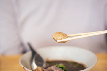 Unrecognizable Asian woman picking up a meat ball from the noodle bowl by using a wooden chopsticks close up. 