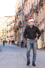 Uomo con cappello da babbo natale, camicia, jeans e mascherina chirurgica,  in centro città tra le vetrine dei negozi