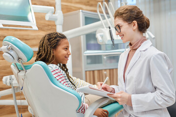 Obraz premium Young female dentist talking to her little patient and making notes in medical card at dental clinic