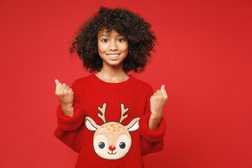 Little african american curly kid girl 12-13 years old wearing knitted cozy deer Christmas sweater doing winner gesture isolated on red background children studio portrait Childhood lifestyle concept