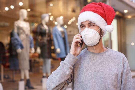 Uomo con cappello da babbo natale, maglia grigia e mascherina ffp2 parla al cellulare mentre passeggia in centro tra le vetrine dei negozi