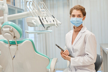 Portrait of young dentist in mask and in lab coat holding digital tablet looking at camera while standing at office