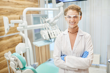 Portrait of young dentist in white coat and in protective glasses standing with arms crossed and smiling at camera in clinic
