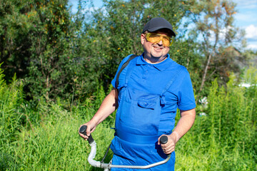 Close-up, funny fat man in blue overalls and a baseball cap, mowing the grass with a trimmer.