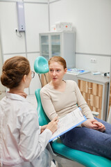 Young woman sitting on dental chair and talking to dentist while she filling the medical card after medical procedure at clinic