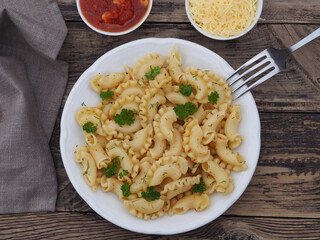 Curly pasta, macaroni with spices and herbs in a plate, tomato sauce, grated parmesan cheese, fork on a wooden table, closeup, top view. Simple delicious rustic dish with pasta products