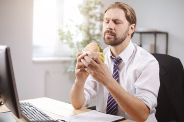 Handsome man in white suit and tie sitting at modern office and eating burger during break. Bearded businessman having lunch with fast food at workplace.