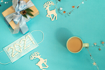 Flat lay of blue working table background with cup of coffee and Christmas decoration: gift box, deer, mask with pills in form number 2021