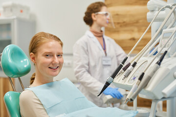 Portrait of young patient smiling at camera while sitting on dental chair with dentist in the background