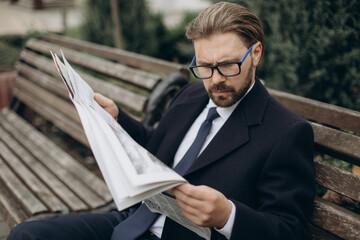 Stylish bearded man in eyewear reading newspaper while sitting on bench at city street. Concept of business, news and information.