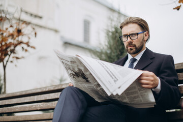 Focused businessman in elegant suit and eyewear reading daily news during free time on fresh air. Bearded manager sitting on bench and holding newspaper.