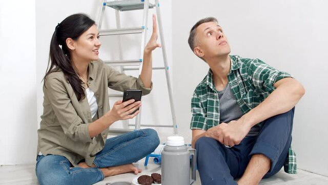 Young happy couple using smartphone while shopping online or getting inspired by new ideas for renovation sitting on the floor in the room. New housing, online shopping and renovation concept