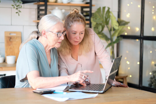 Senior woman using laptop for websurfing in her kitchen. middle-aged daughter helps her mother with documents. Mature lady sitting at work typing a notebook computer in an home office.