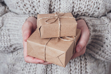 Gifts in women's hands, close-up. Monochrome, minimalist gift concept. A girl in a sweater holds gift boxes made of Kraft paper, tied with a string. Surprise background, greeting card