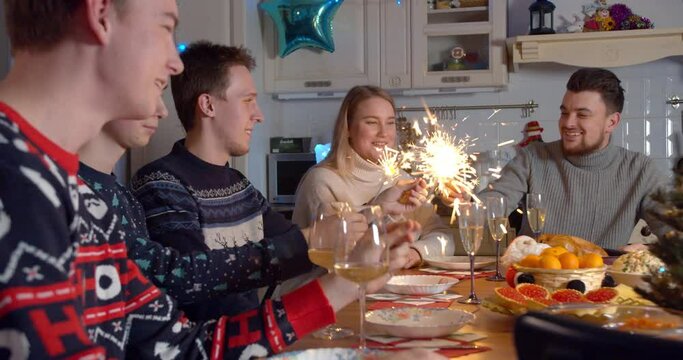Friends in sweaters are drinking champagne talking on Christmas Eve 2021 with sparklers, lights. Happy people celebrating new year together on quarantine covid-19 coronavirus concept. 