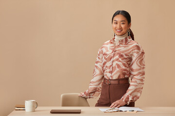 Fototapeta na wymiar Portrait of Asian young businesswoman smiling at camera standing near her workplace against the beige background