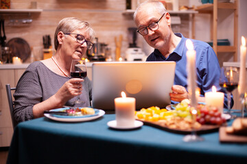 Aged man and woman shopping online using laptop in kitchen during festive dinner. Woman holding glass of wine. Elderly people sitting at the table browsing, searching, using laptop, technology