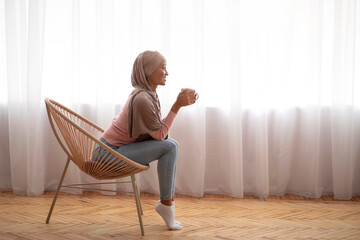 Fototapeta premium Side view of black Muslim woman in hijab drinking hot coffee in cozy chair near window indoors, free space