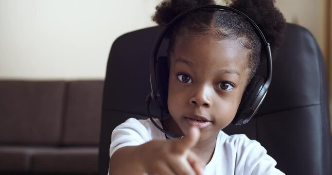 Portrait of small emotional African girl 5 -7 years old sits at table at home in front of computer laptop wears head microphone speaks communicates online studies remotely points to camera with finger