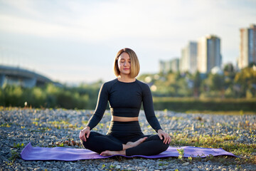 Fototapeta na wymiar calm young caucasian woman is meditating in lotus pose outdoors, sit with eyes closed, alone on fresh air, wearing sportswear