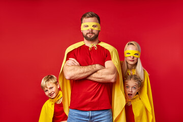 cute mother and kis hide behind a super man's back wearing yellow cloaks and masks, costume of...