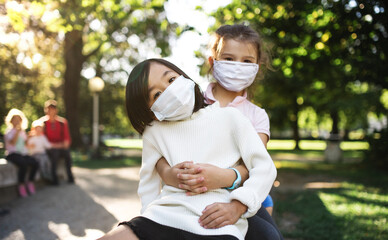 Small school girls with face mask on playground outdoors in town, coronavirus concept.