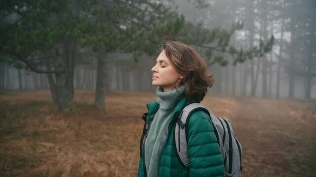 A cinematic shot of a young cheerful woman makes a deep breath and enjoy the fresh air while walking in the misty forest on a fall day.