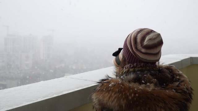 A warm-dressed woman drinks coffee and looks at the falling snow flakes.