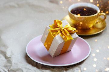Obraz na płótnie Canvas Zero gravity gift box wrapped by kraft paper with yellow satin ribbon on pink handmade plate. Gold coffee cup unfocused. Universal gifts design for all kind of celebration and sales